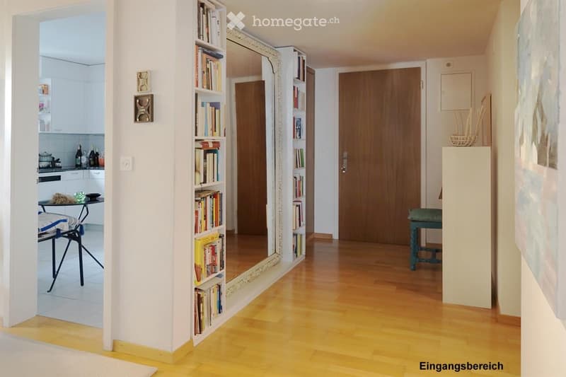 Bright hallway with wooden flooring, large mirror, bookshelf, and a small table with a chair in the adjacent kitchen area.