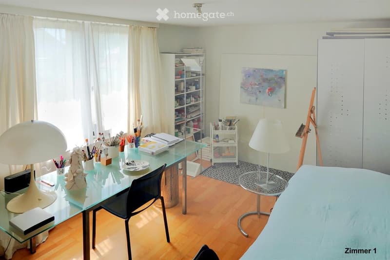 Well-lit room with a glass desk, chair, two lamps, a cabinet, a painting on the wall, a small stool, and an art easel. The room features wooden flooring and large windows with curtains.
