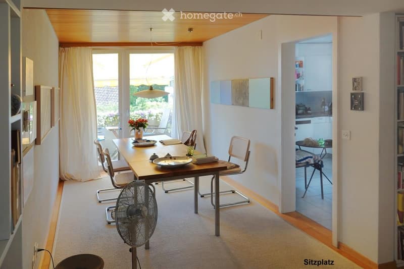 Bright, open-plan living and dining area with a wooden dining table, chairs, a fan, and a view to a kitchen area with cabinets and a small table.