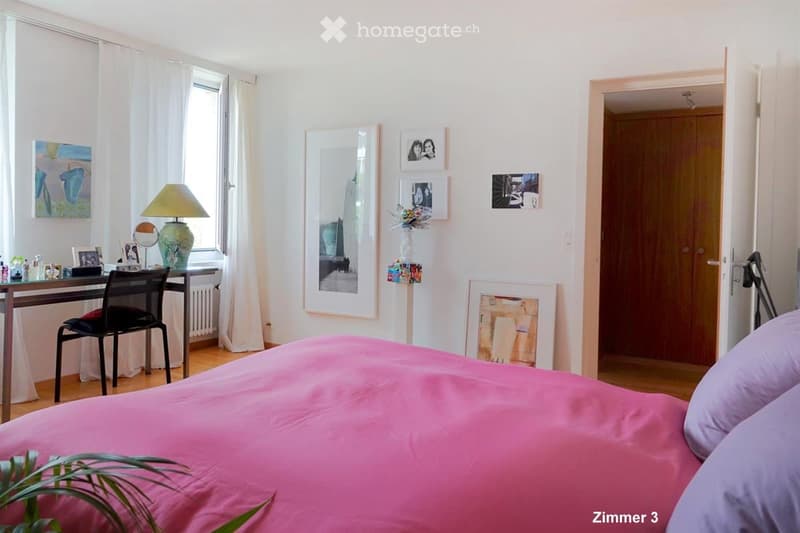Bright bedroom with a pink bedspread, a wooden desk with a chair, a lamp, and several picture frames on the wall.