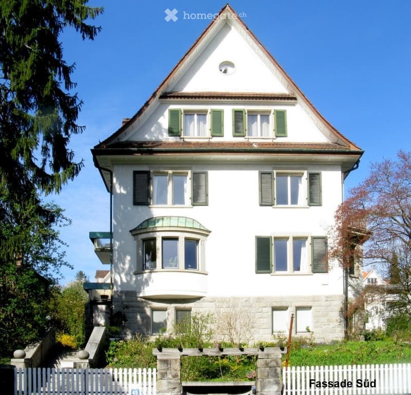 Two-story white house with green shutters, a bay window, and a sloped roof. The house is surrounded by a small garden with a white picket fence.