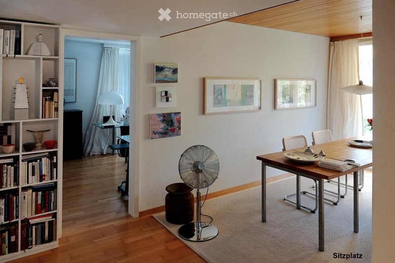 Bright, modern room with wooden flooring, large window with white curtains, a round wooden table with chairs, a standing fan, a bookshelf filled with books and decorative items, and several framed artworks on the wall.