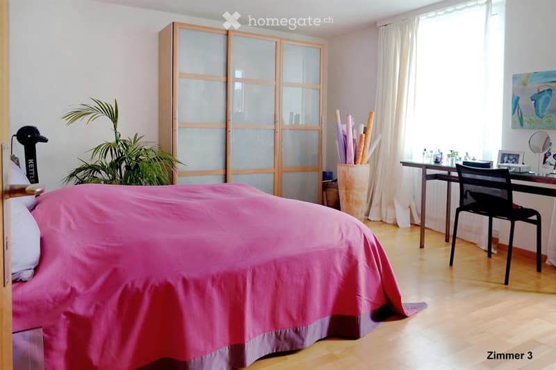 Bright bedroom with wooden floor, large bed with pink bedding, a wardrobe with glass doors, a desk with chair, a plant, and a mirror on the wall.