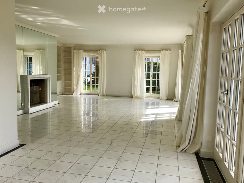 Empty room with white tiled flooring, large windows with white curtains, and a fireplace on the left side.