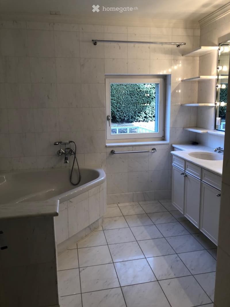 Bathroom with a bathtub, sink, and cabinet, featuring tiled walls and floor, a window with a view of greenery and a pool.