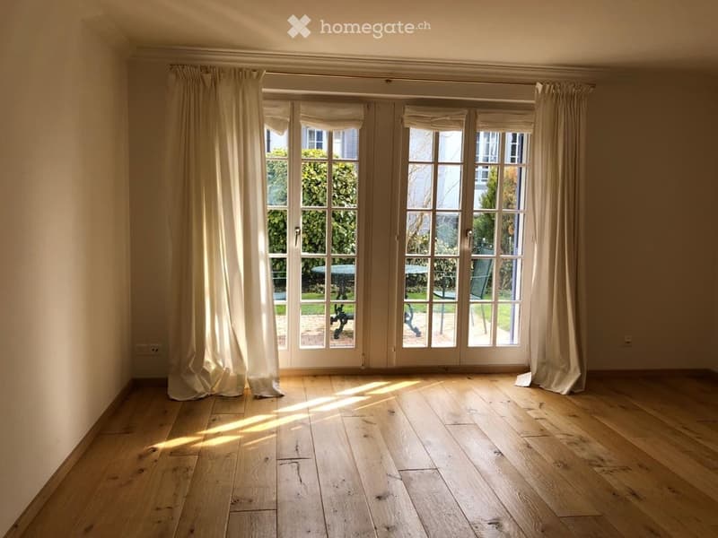 Empty room with wooden flooring and large glass doors leading to an outdoor area, with light-colored curtains on either side.