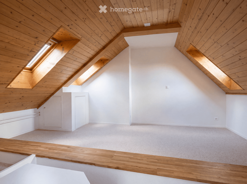 Empty attic room with wooden slanted ceilings, skylights, white walls, and a wooden floor border.