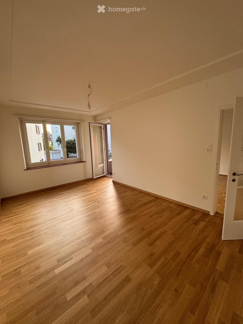 Empty room with wooden flooring, white walls, large window, and open door leading to another space.