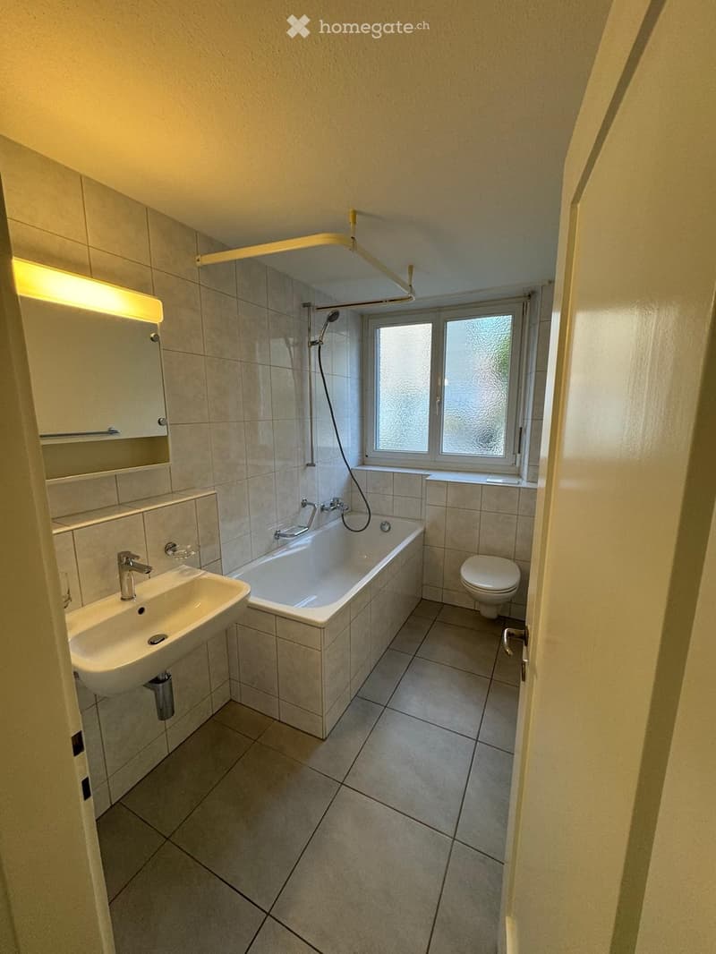 Modern bathroom with a bathtub, sink, toilet, and mirror. Features tiled walls and floor, a window with frosted glass, and a showerhead above the bathtub.