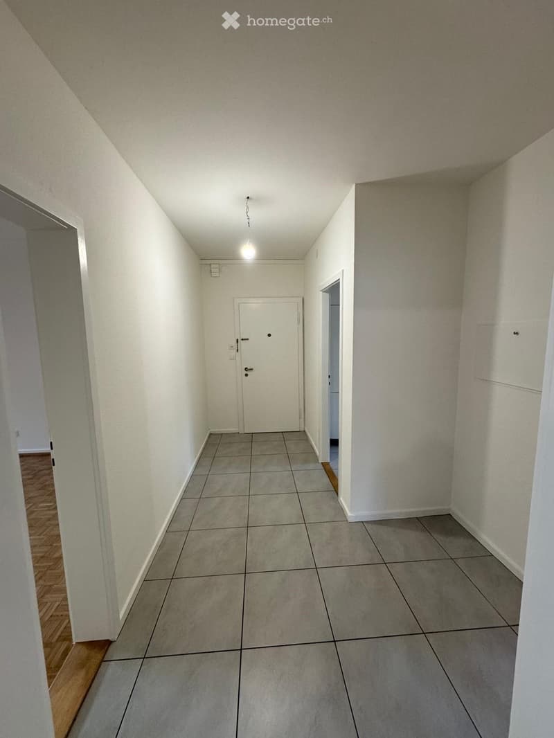 Empty hallway with tiled floor, white walls, and a single door at the end. The ceiling has a light fixture, and there are visible doorways on both sides.