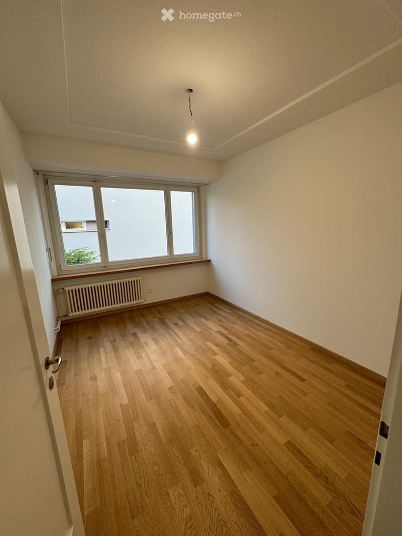 Empty room with light wooden flooring, white walls, and large windows with a radiator below. No furniture or personal items are present.