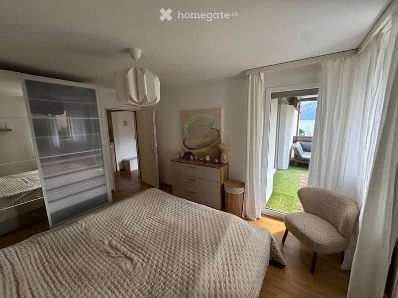 Cozy bedroom with a large bed, a modern chair, a dresser, a mirror, and a large window with white curtains leading to a small outdoor area.