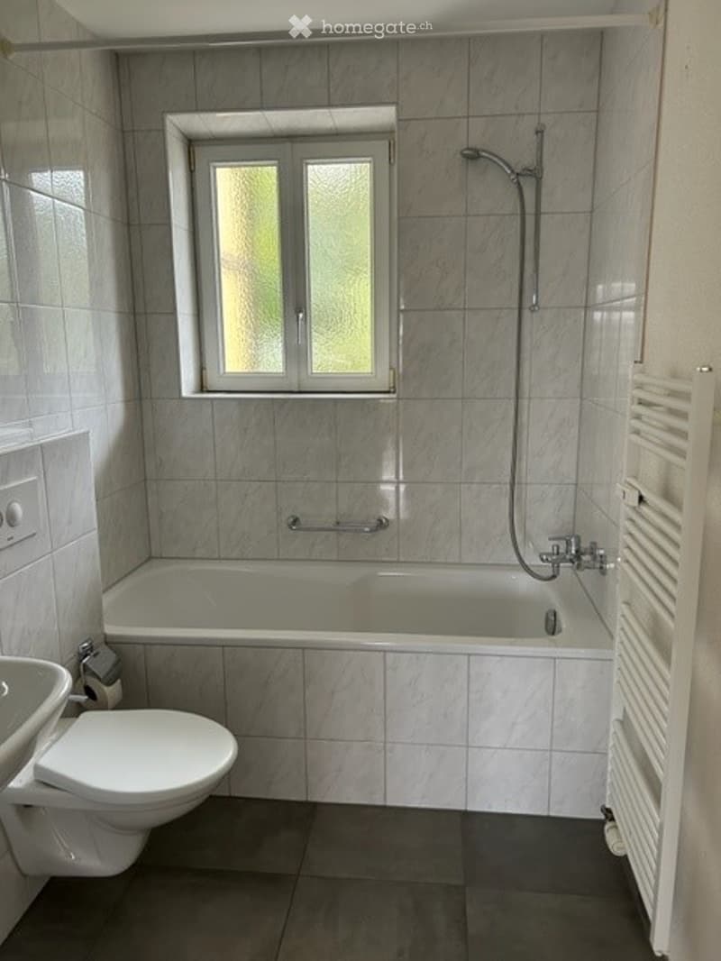 Modern bathroom with a bathtub, toilet, and showerhead, featuring tiled walls and a window for natural light.