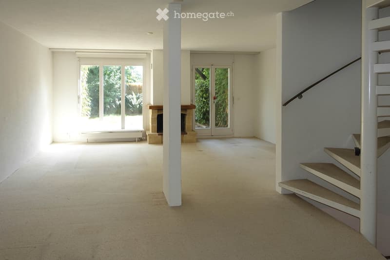 Empty, unfurnished room with white walls, carpeted floor, and a staircase leading upwards. Features a fireplace and large windows with a view of greenery.