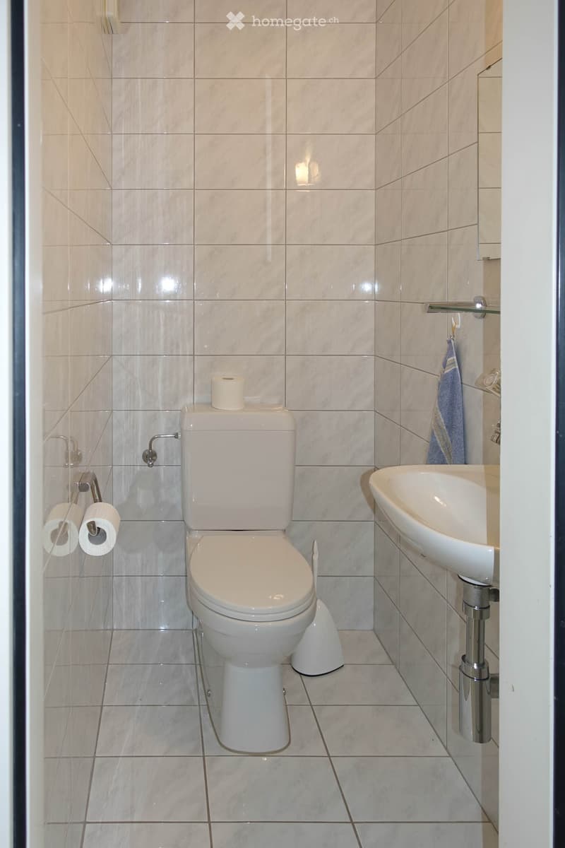 Modern bathroom with white tiles, toilet, sink, and two rolls of toilet paper on a holder. A blue towel hangs on the wall.