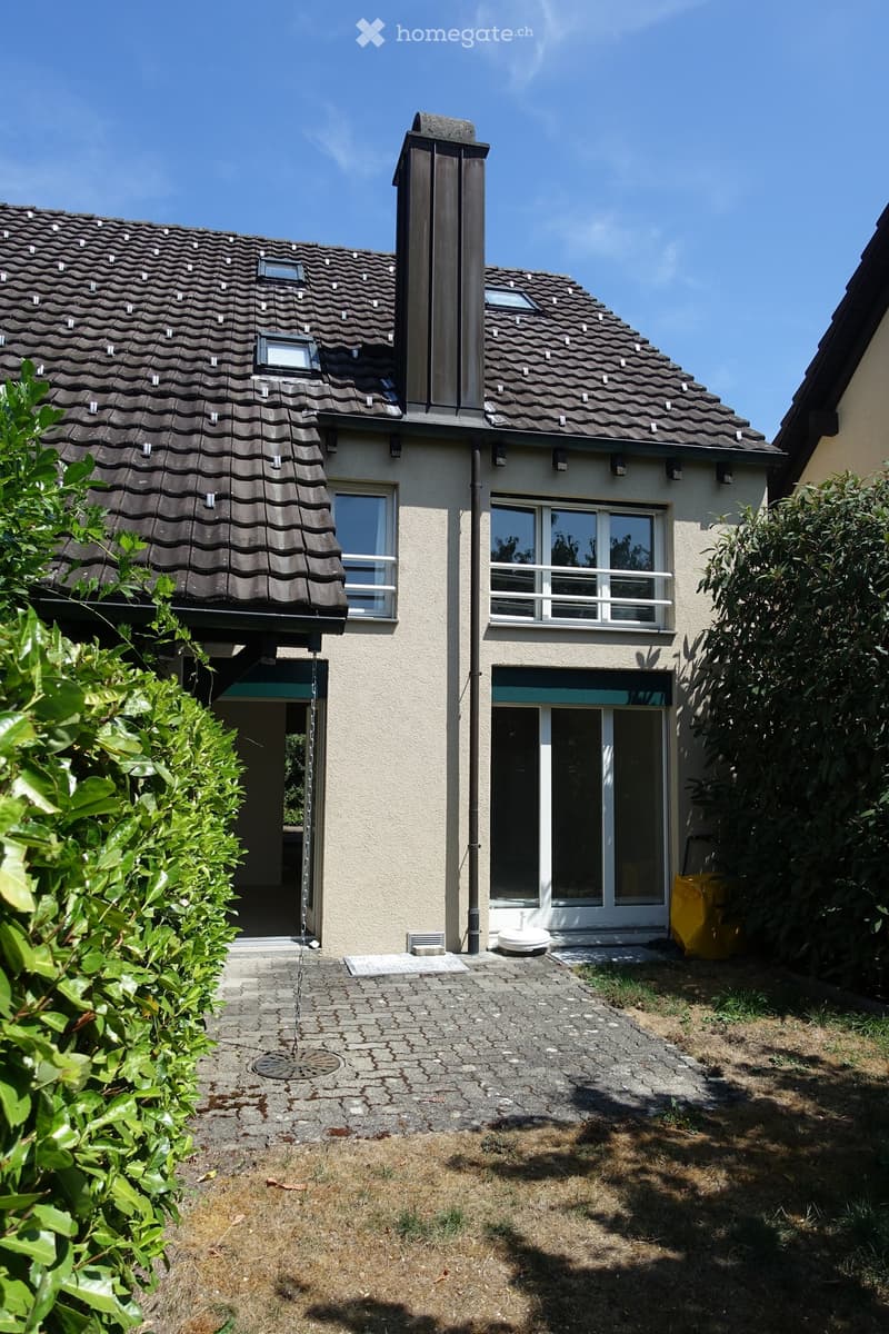 Two-story house with a tiled roof, multiple windows, and a small front yard with a paved pathway leading to the entrance.