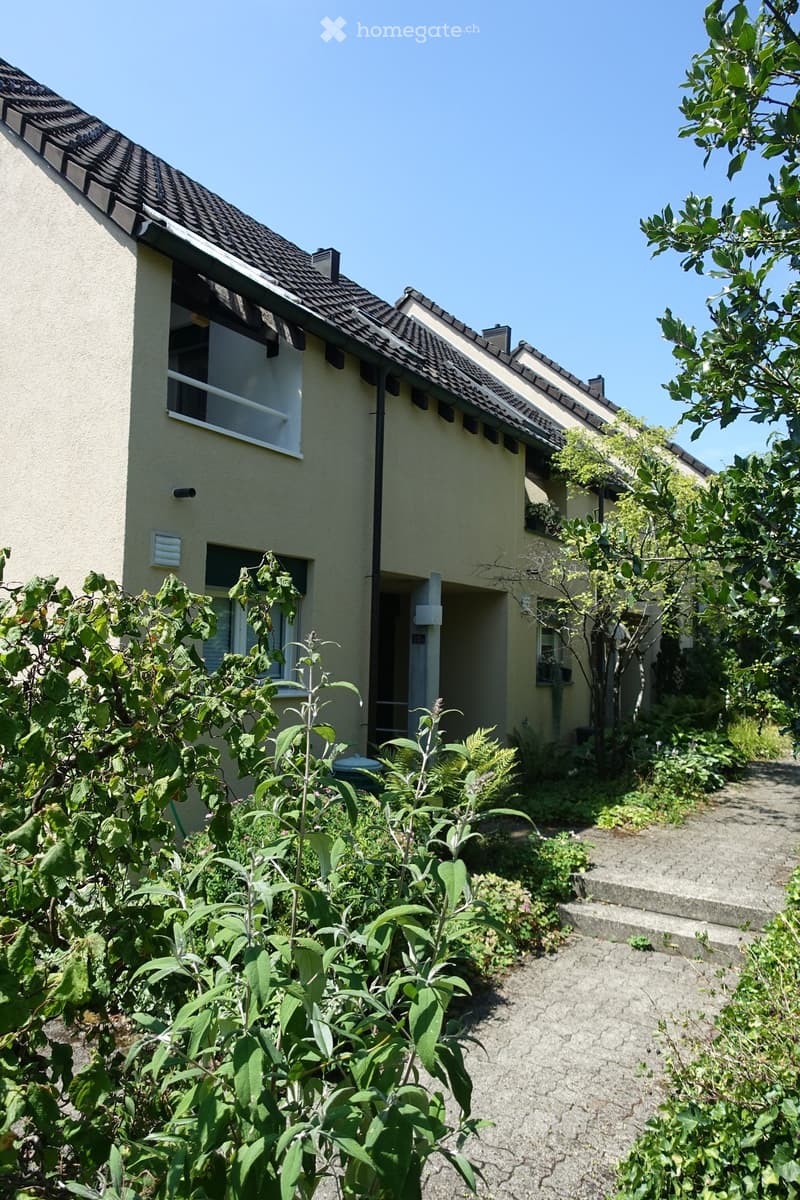 Two-story house with beige exterior, dark brown tiled roof, multiple windows, and a small garden area with various plants and trees.