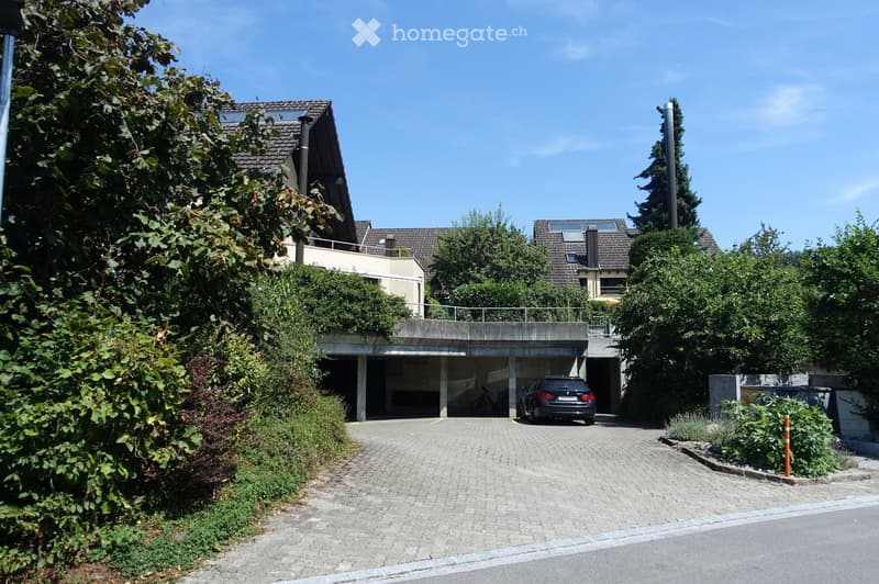 Modern multi-story building with a covered garage area, surrounded by greenery and trees.