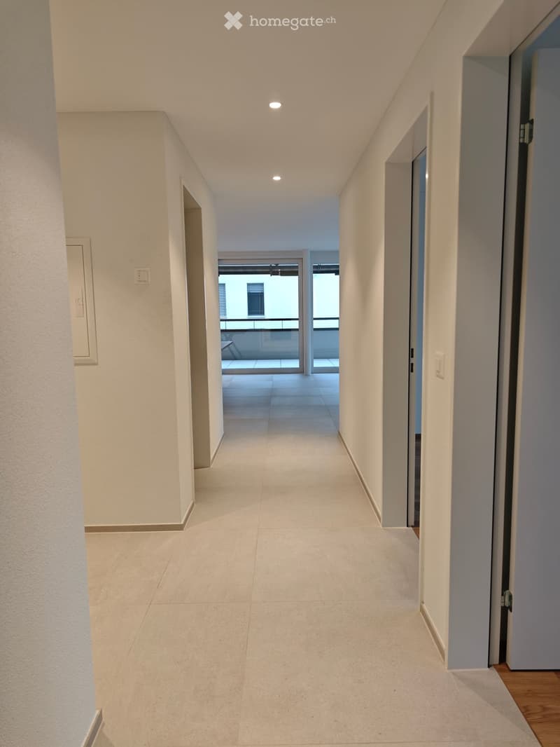 Modern hallway with light-colored tiled flooring, white walls, and recessed lighting. Features a door leading to a balcony area with a view of the exterior.