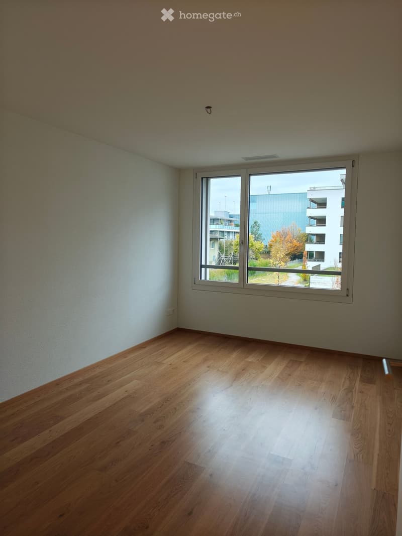 Empty room with wooden flooring and large window showing view of buildings and trees outside.