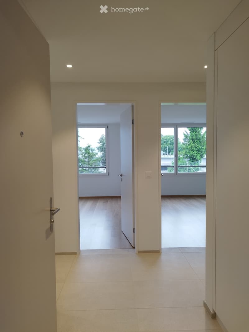 Empty hallway with tiled floor, white walls, and two open doors leading to rooms with wooden flooring and large windows offering a view of greenery.