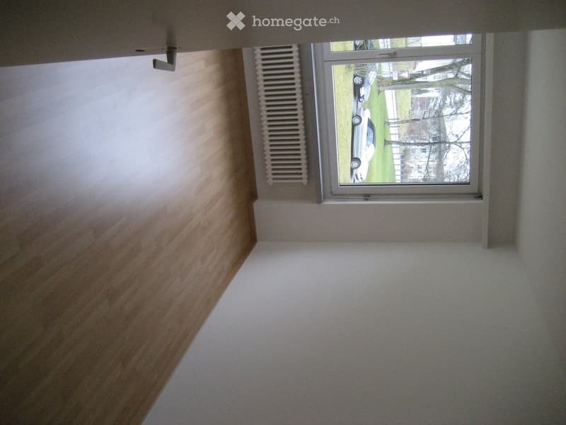 Empty room with light-colored walls, wooden paneling on one wall, and a window showing a view of a car and outdoor area.