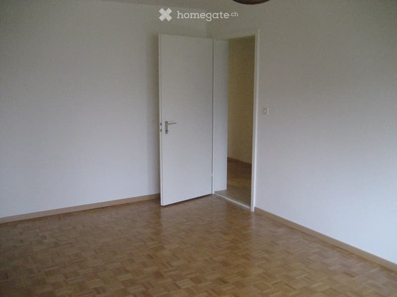 Empty room with wooden flooring and white walls, featuring a closed white door and a partially visible adjacent space.