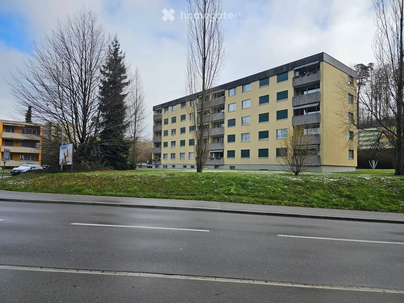 Multi-story residential building with yellow facade, multiple windows, and balconies, surrounded by trees and a grassy area.