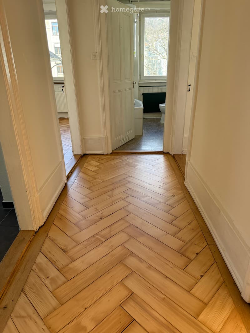 Hallway with herringbone-patterned wooden flooring, white walls, and doors leading to other rooms.