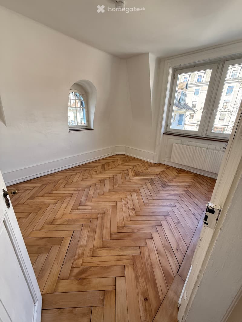 Empty room with herringbone wooden flooring, white walls, and large windows offering a view of the city.