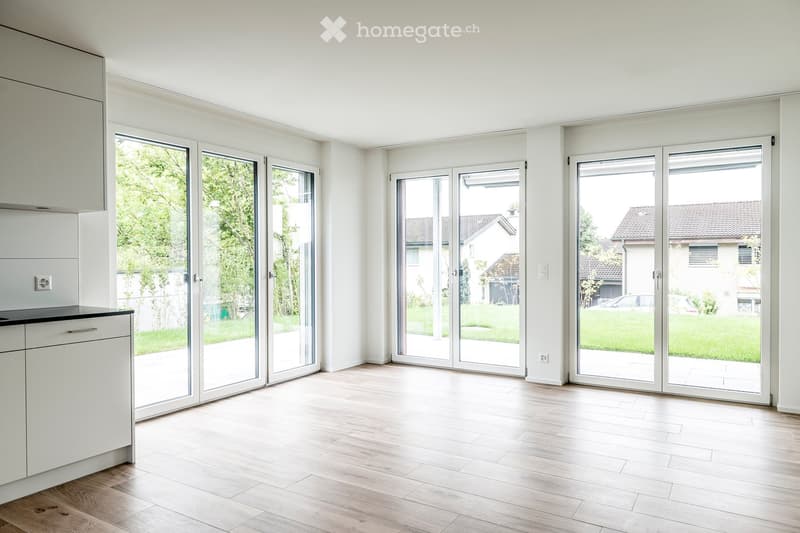 Bright, unfurnished room with light wooden flooring, white walls, and large sliding glass doors leading to a garden. Minimalist design with a small cabinet visible on the left side.