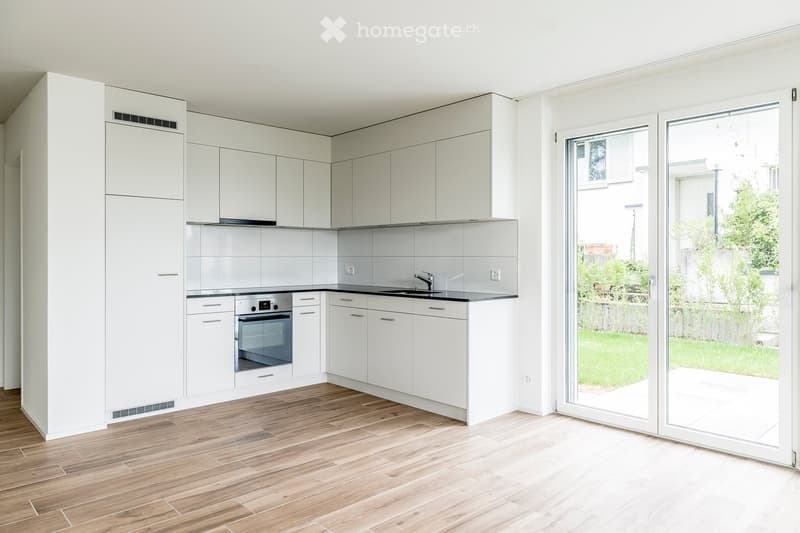 Modern, minimalist kitchen with white cabinets, wooden flooring, and a large sliding glass door leading to a garden.