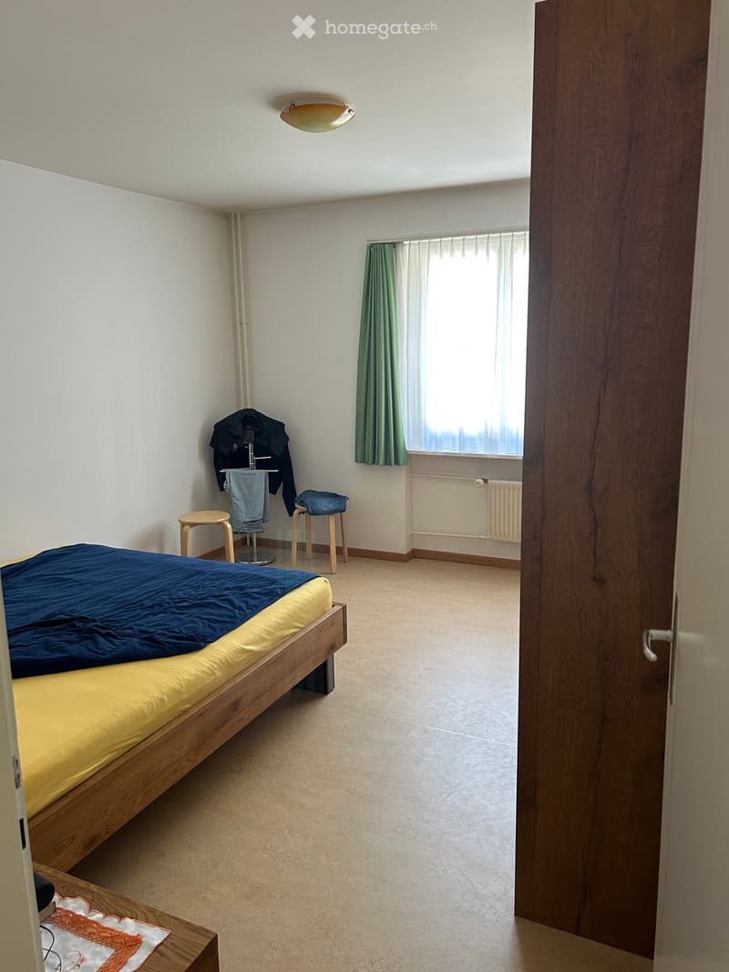 A small, furnished bedroom with a wooden bed, two wooden stools, a radiator, green curtains, and a wardrobe with clothes.
