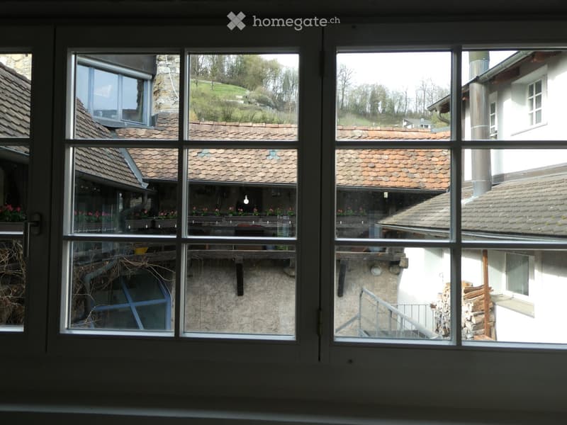 View from a window showing a rural landscape with traditional houses, tiled roofs, and a stone wall with stacked firewood.
