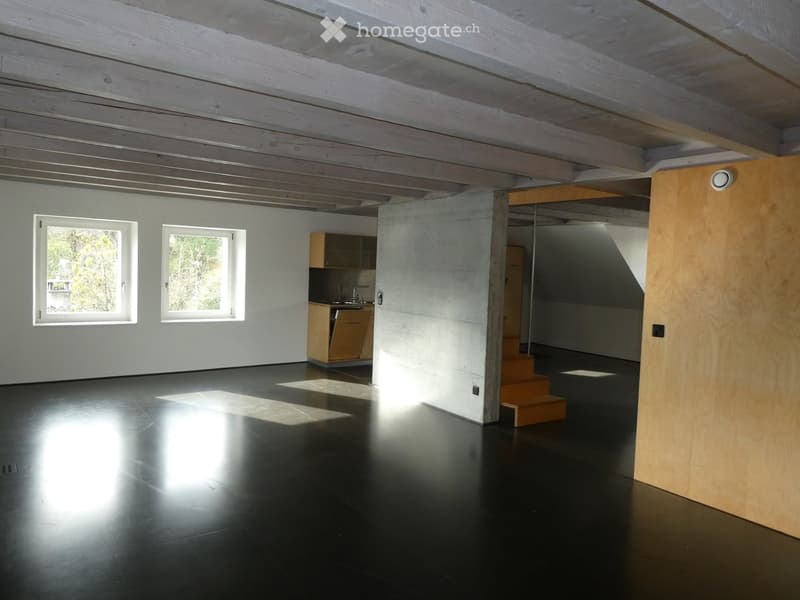 Open-plan space with dark flooring, exposed wooden ceiling beams, two windows, small kitchen area with cabinets and sink, and a wooden staircase leading upwards.