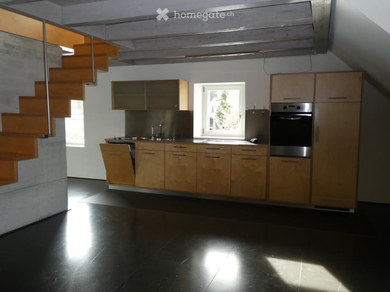 Modern kitchen with light wooden cabinets, stainless steel appliances, and a black tiled floor. Features a window, sink, and a wooden staircase leading upwards.