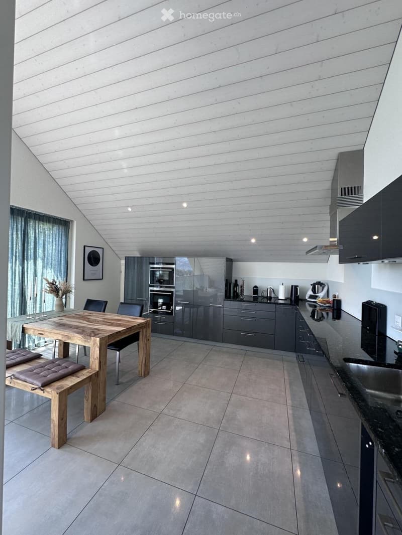 Modern kitchen with grey cabinets, wooden dining table, and seating area. Features include an oven, refrigerator, and a coffee maker on the counter.