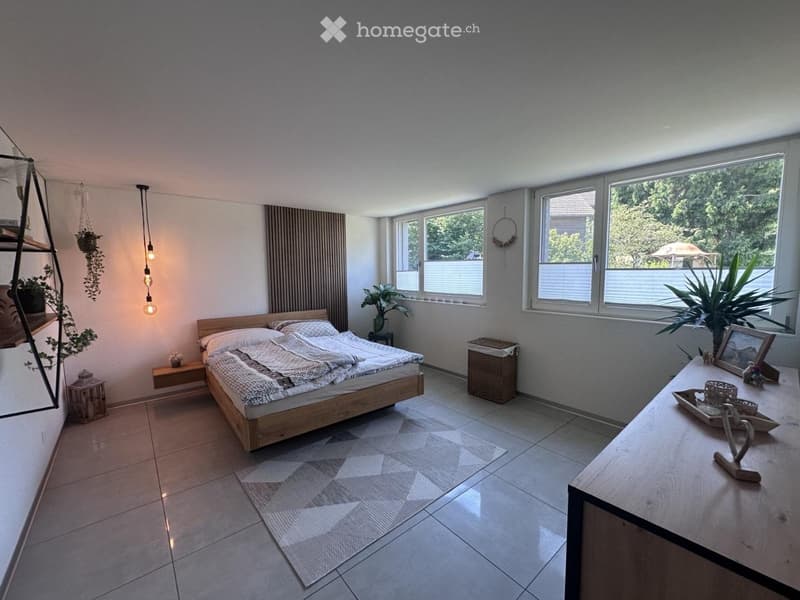 Modern bedroom with a wooden bed, nightstand, side table, and decorative plants. Large windows provide natural light.