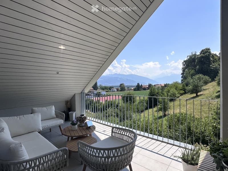 Balcony with a view of the countryside and mountains, featuring a sofa, a round table, and two wicker chairs.