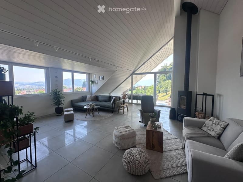 Modern living room with large windows offering a scenic view, featuring a grey sectional sofa, coffee tables, ottomans, armchairs, and a fireplace.
