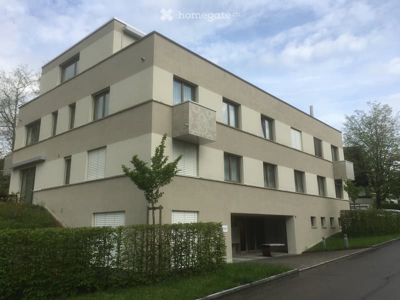Modern two-story house with beige facade, large windows, and a small balcony on the upper floor. The house features a raised ground floor, a garage entrance, and is surrounded by greenery and a small tree.