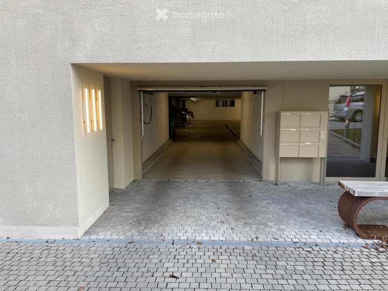 Entrance to a covered parking area with a concrete floor, beige walls, and a small seating area to the right.