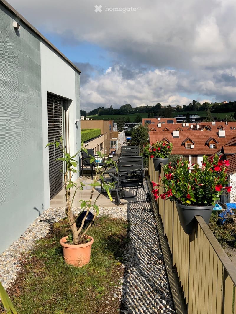 Balcony with a small seating area, potted plants, and a view of the surrounding neighborhood.