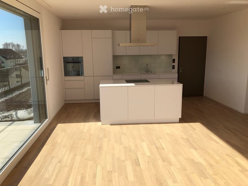 Modern, minimalist kitchen with white cabinetry, a central island, and a range hood. The space features light wooden flooring and large windows offering a view of the outdoor area.