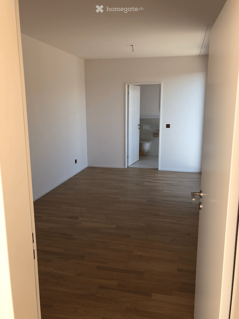 Empty room with wooden flooring and white walls, featuring an open doorway leading to a small bathroom with a toilet.