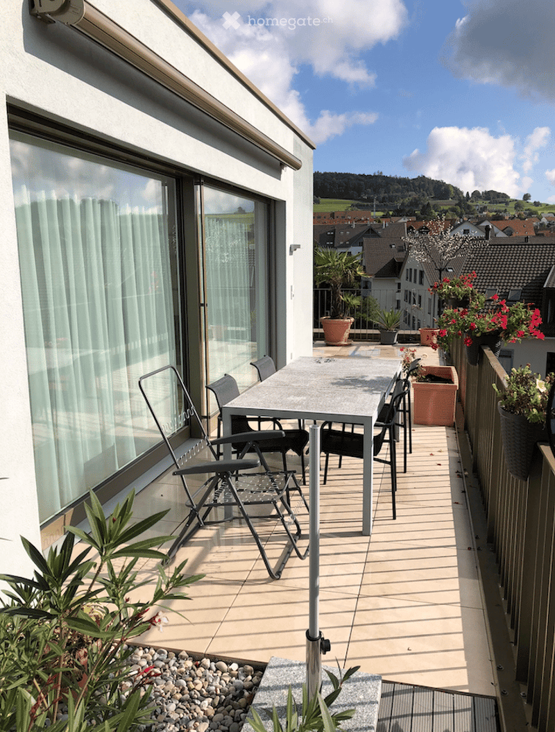 Spacious balcony with a modern dining table and chairs, surrounded by potted plants and a scenic view of the town and hills.