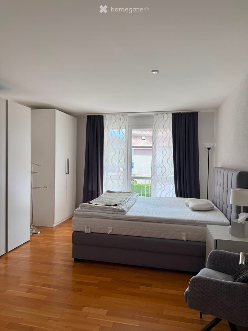 Modern bedroom with wooden flooring, large window with dark curtains, grey upholstered bed with headboard, nightstand with lamp, grey armchair, and white wardrobe.