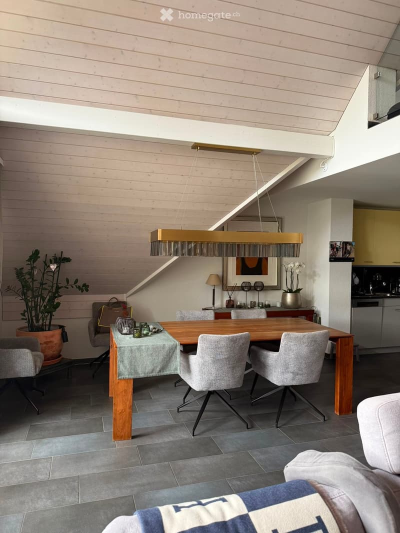 Modern dining area with a wooden dining table, six chairs, a sofa, pendant lighting, and a potted plant. The space features a sleek kitchen area with cabinets and a countertop.