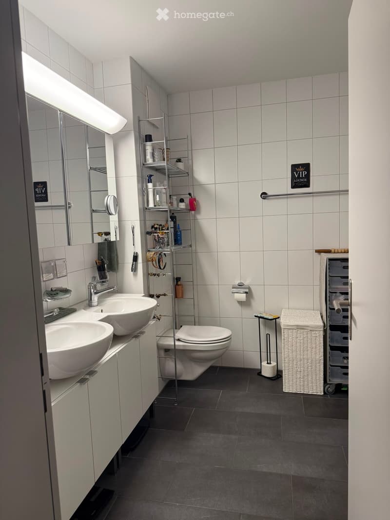 Modern bathroom with two sinks, a toilet, a mirror, a towel rack, a storage shelf, and a wicker basket. The walls are tiled, and the floor is dark grey.