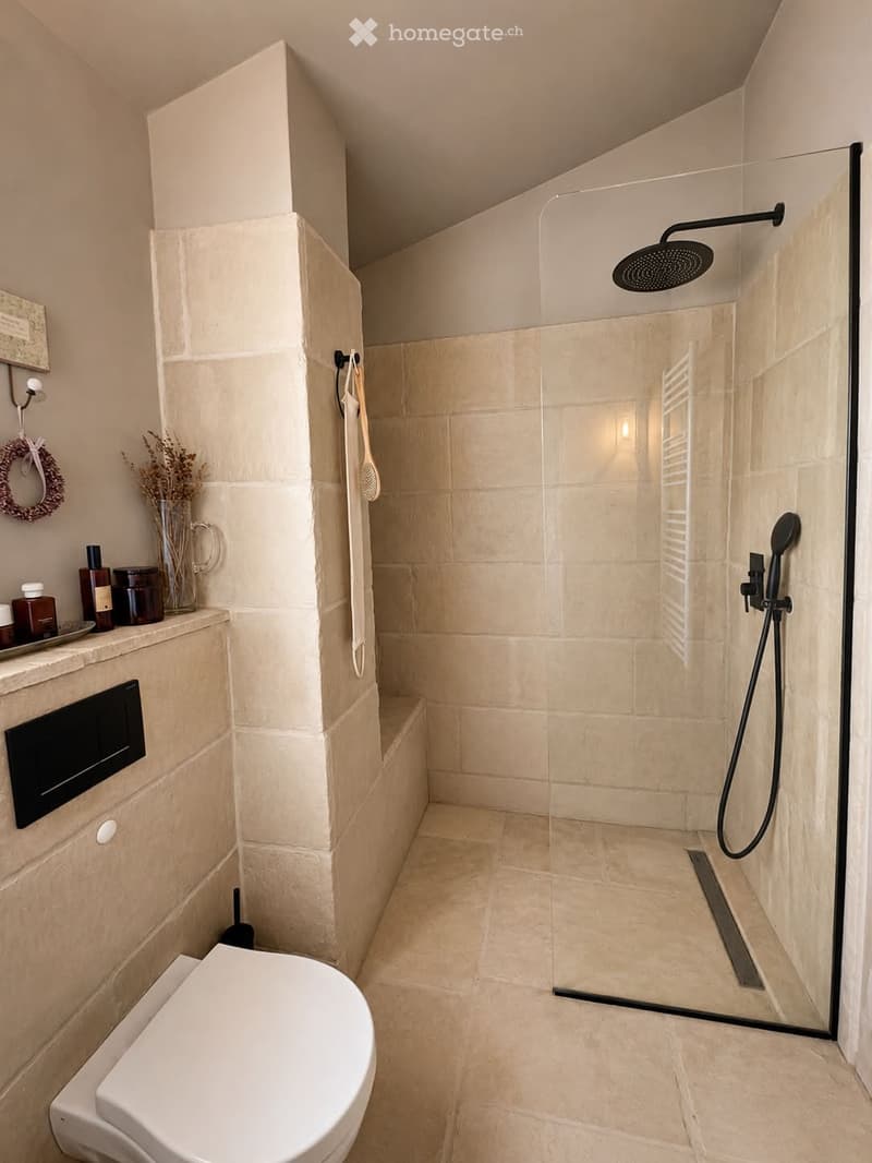 Modern bathroom with a large walk-in shower, toilet, and a variety of toiletries on the counter. The space features beige tiles and a sleek glass shower enclosure.